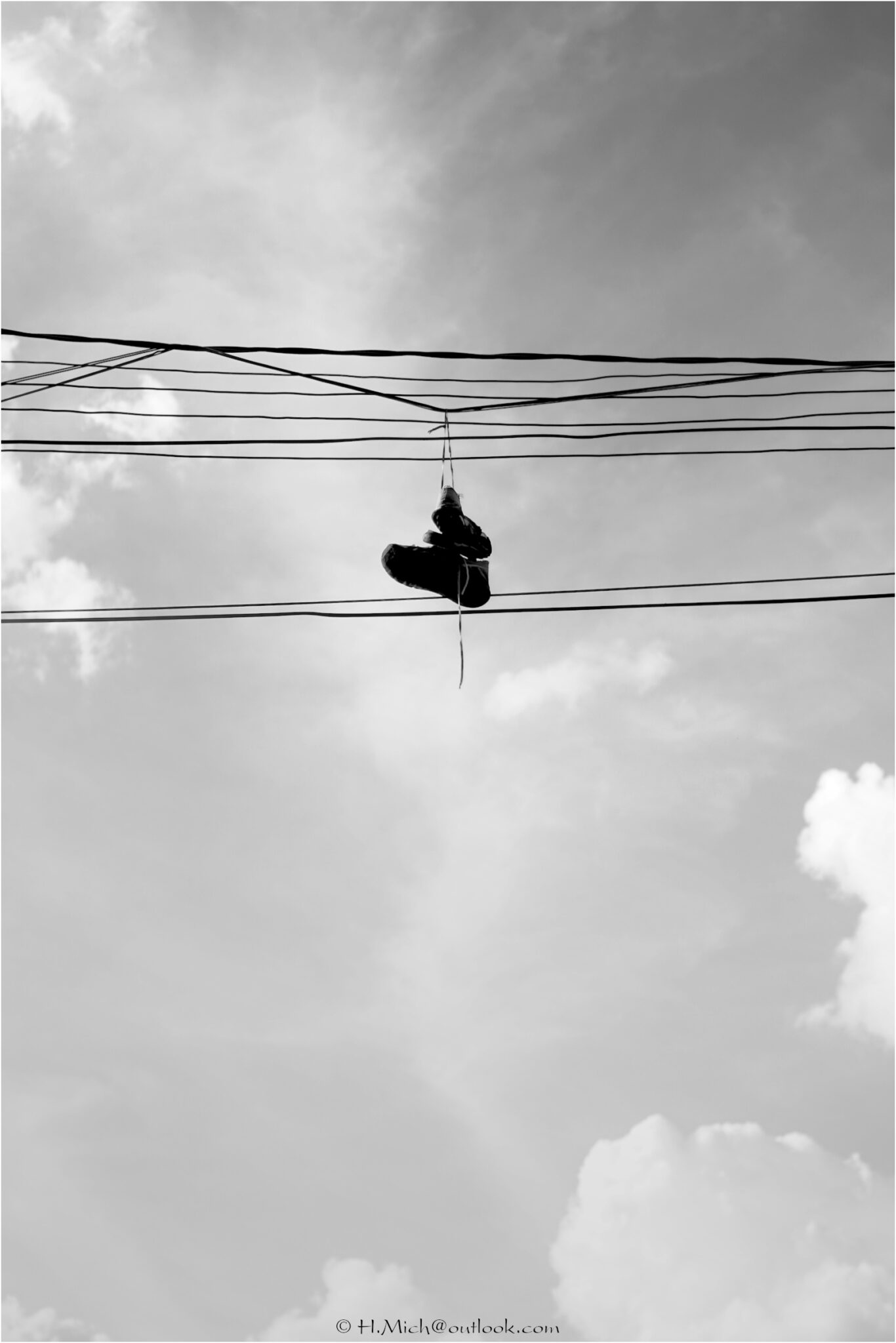 Diarium.Studio-One.Art.Br - Um par de tênis pendurado pelos cadarços em linhas de energia aéreas contra um céu nublado. A fotografia em preto e branco captura os sapatos suspensos no ar, com várias linhas paralelas visíveis, transmitindo uma sensação de altura e isolamento. - (c) H.Mich
