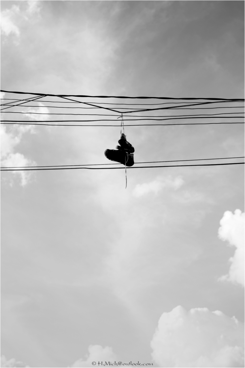 Diarium.Studio-One.Art.Br - Um par de tênis pendurado pelos cadarços em linhas de energia aéreas contra um céu nublado. A fotografia em preto e branco captura os sapatos suspensos no ar, com várias linhas paralelas visíveis, transmitindo uma sensação de altura e isolamento. - (c) H.Mich
