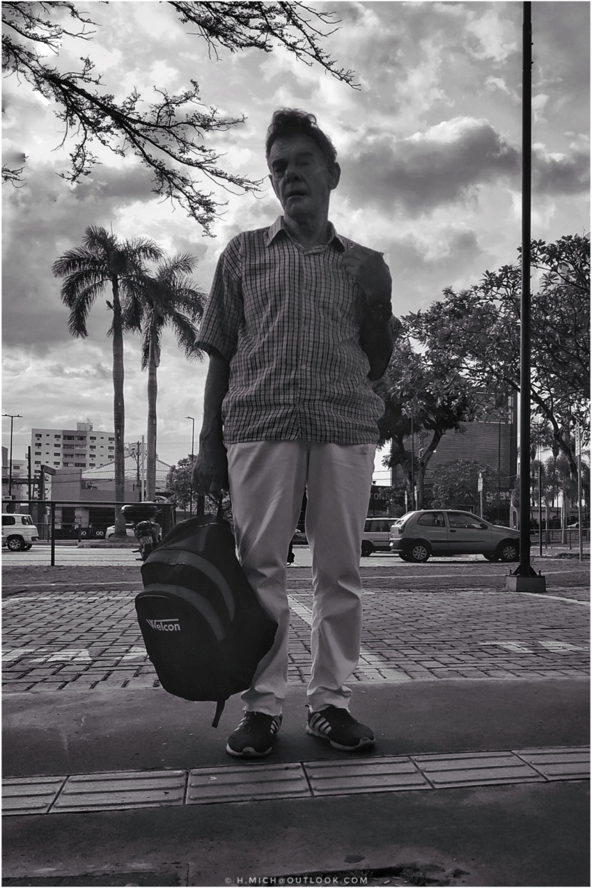 Diarium.Studio-One.Art.Br - Foto em preto e branco de um homem parado na calçada, segurando uma mochila com WAGON escrito nela. Ele veste uma camisa xadrez, calças leves e tênis. Árvores e uma paisagem urbana estão ao fundo, com carros e prédios sob um céu nublado. - (c) H.Mich