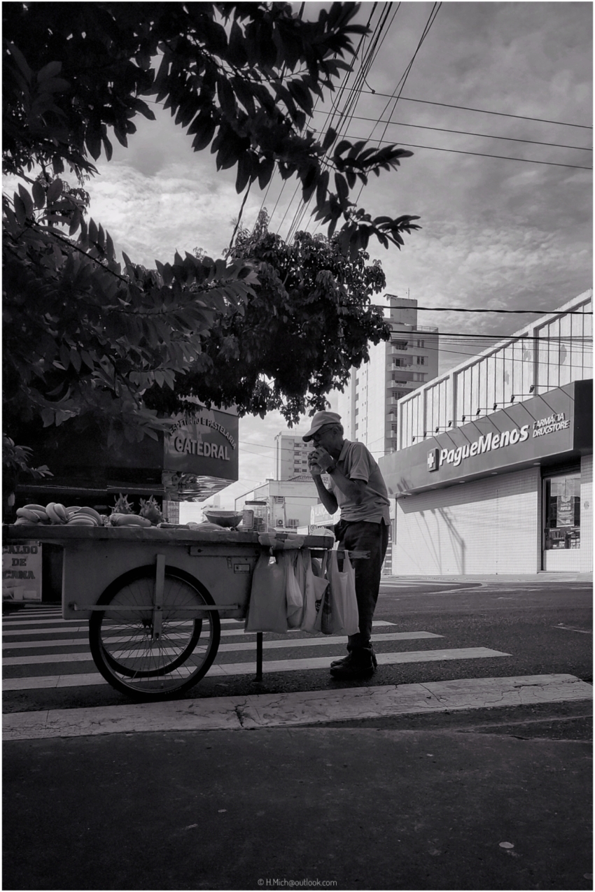 Diarium.Studio-One.Art.Br - Homem usando chapéu e em pé ao lado de um carrinho de rua cheio de produtos e salgadinhos, bebendo de um copo. O carrinho está posicionado em uma faixa de pedestres em uma rua da cidade. Prédios estão ao fundo, incluindo uma farmácia Pague Menos. Fios aéreos e galhos frondosos são visíveis. Foto em preto e branco. - (c) H.Mich