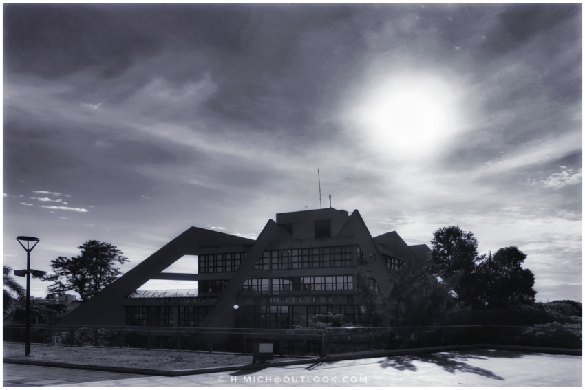 Diarium.Studio-One.Art.Br - Foto em preto e branco de um edifício moderno e geométrico com telhados angulares e janelas grandes. O sol brilha intensamente no céu, parcialmente obscurecido pelas nuvens. Um poste de luz solitário fica à esquerda, e árvores emolduram o edifício. O primeiro plano apresenta uma área pavimentada. - (c) H.Mich